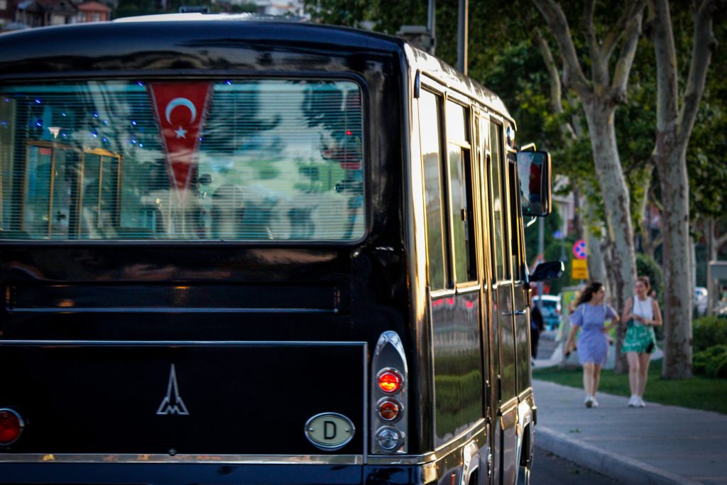 A bus with a Turkish flag in the city, two women walking nearby on a sunny day.